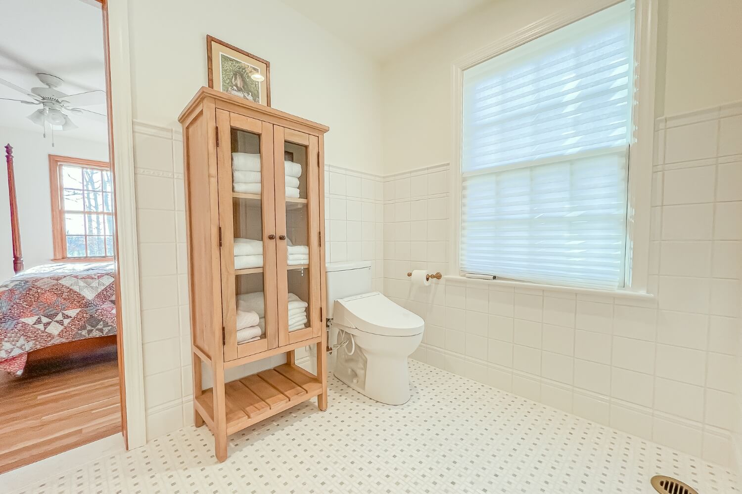 A stylish wooden storage cabinet in a white tiled bathroom, with a glimpse of a bedroom.