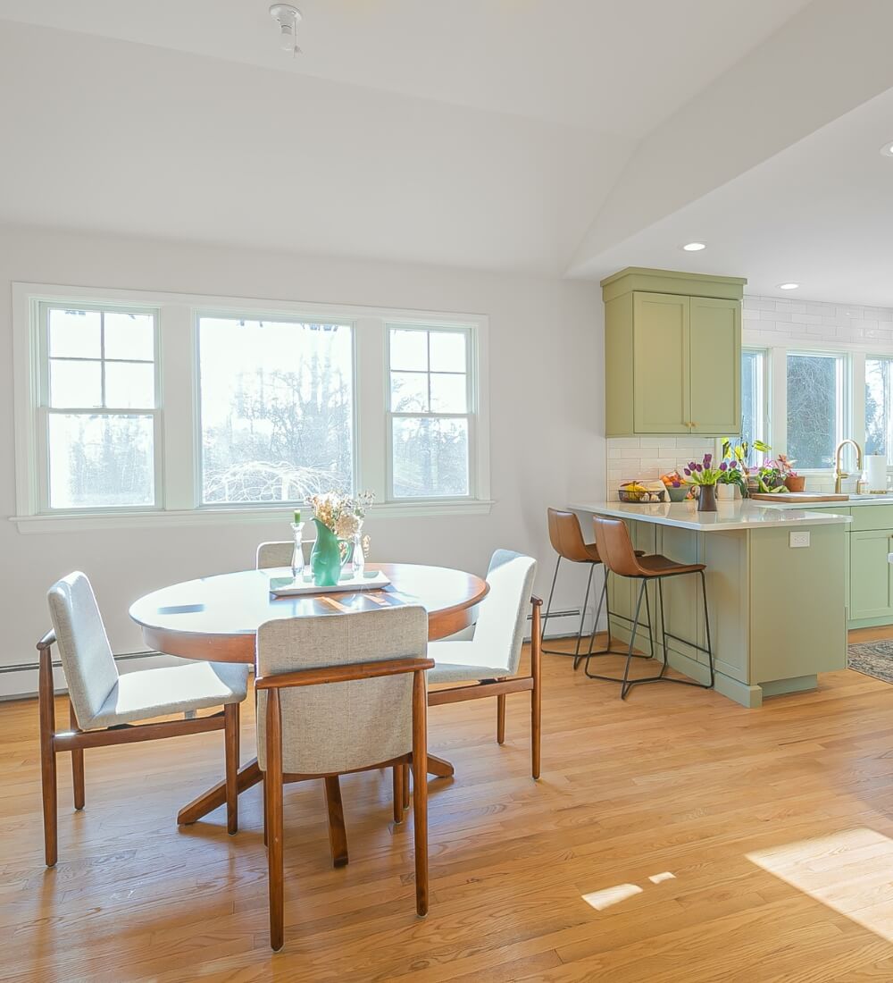 A spacious, sunlit open-concept kitchen featuring sage green cabinets, a white countertop island with barstools, and a cozy wooden dining table with upholstered chairs.